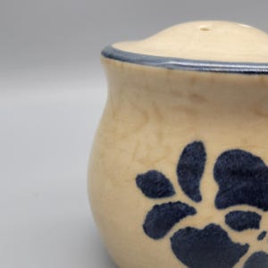 May include: Cream-colored ceramic salt and pepper shakers with a blue floral design. The shakers have a rounded shape with a lid and a blue trim. The floral design is a dark blue color.