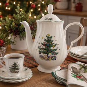 May include: A white porcelain coffee pot and matching teacup with saucer, featuring a festive Christmas tree graphic with colorful ornaments, a star topper, and wrapped gifts at the base, accented by a delicate green rim and fluted texture.