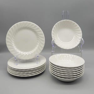May include: A set of white ceramic dinnerware, including plates and bowls. The plates and bowls have a spiral design around the edges. The dinnerware is displayed on clear plastic stands against a gray background.