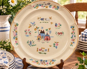 Può includere: Un piatto piano rotondo in ceramica con base color crema, caratterizzato da un'illustrazione centrale di case, animali da fattoria e una persona che fa giardinaggio. Il bordo è decorato con un motivo floreale colorato e un uccello.
