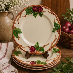 May include: A stack of cream-colored ceramic plates with a decorative apple and branch design. The plates have a brown rim and are supported by a wooden plate stand. A basket of red apples and a vase of white flowers are in the background.