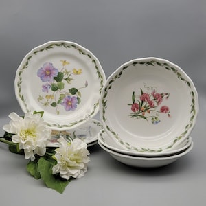 May include: A set of white ceramic plates and bowls with floral designs. The plates and bowls have a scalloped edge with a green leaf border. The floral designs include purple, pink, and yellow flowers.