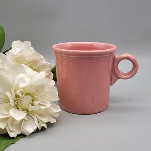 May include: A pink ceramic mug with a rounded handle. The mug has a smooth, glossy finish and a simple, classic design. The mug is a solid pink color and appears to be a standard size for coffee or tea.