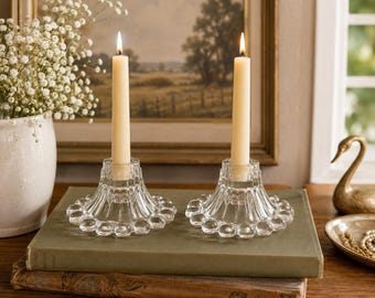 Puede incluir: Dos velas de color marfil encendidas en candelabros de cristal transparente con borde festoneado. Los candelabros están sobre una pila de libros. Un jarrón con flores blancas y una pintura de paisaje enmarcada están en el fondo.