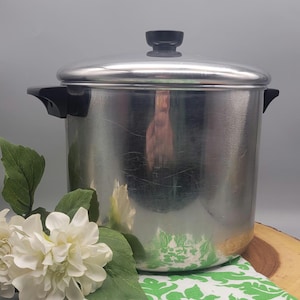 May include: A stainless steel cooking pot with a lid and black handles. The pot is sitting on a wooden surface with a green and white patterned cloth and white flowers.