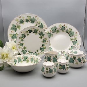 May include: A complete white dinnerware set featuring a green ivy leaf design. The set includes plates, a bowl, a creamer, a sugar bowl, and salt and pepper shakers. The edges of the plates and bowls have a scalloped design.