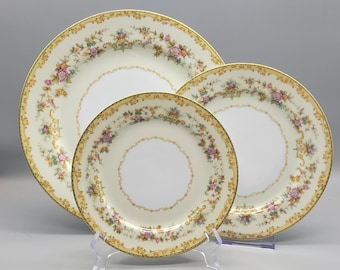 May include: Three round porcelain plates of varying sizes, each featuring a white center, a cream-colored rim, and a decorative border of pink and blue floral garlands with gold filigree accents and a gold-trimmed edge.