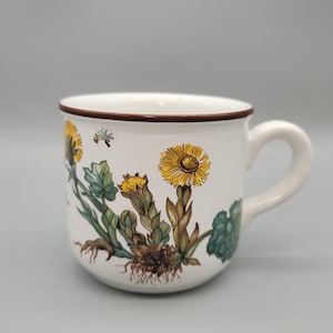 May include: White ceramic mug with a brown rim and handle, decorated with a botanical illustration of yellow flowers, green leaves, and brown roots. A small insect is also depicted. The mug is a standard size for coffee or tea.