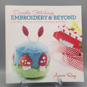 May include: A book titled "Doodle Stitching EMBROIDERY & BEYOND" with a blue, green, and red mushroom design on the cover. The book is propped up on a clear stand. The cover also includes the author's name, Aimee Ray.