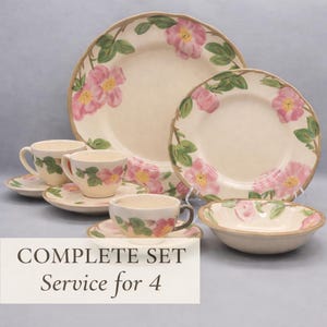 May include: Complete set of cream-colored ceramic dinnerware for four. The set includes plates, bowls, cups, and saucers, all decorated with pink floral and green leaf designs. The rims have a brown trim.