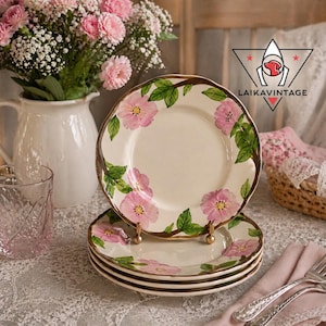 May include: A stack of four cream-colored plates with a gold rim and pink floral design. The plates are stacked on a table with a pink glass, a bouquet of pink flowers, and a pink napkin with silverware.