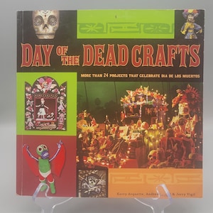 May include: A book titled "Day of the Dead Crafts" with colorful illustrations and the text "More than 24 projects that celebrate Dia de los Muertos." The cover features a skull, figures, and craft projects.
