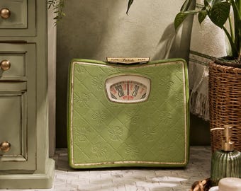 May include: A square bathroom scale in a light green hue featuring a quilted diamond pattern with embossed bow motifs. The scale has a central analog display dial with the text Auto Detect Zero and a white border trim along the edges.