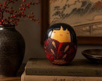 Könnte beinhalten: Eine abgerundete Kokeshi-Holzpuppe mit einem glatten, hellen Gesicht mit einfachen bemalten Zügen, schwarzem Haar und einem dunkelroten Kimono, der mit goldenen und braunen Ahornblattmustern verziert ist, auf zwei gestapelten Büchern.