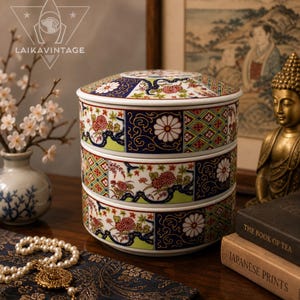May include: A three-tiered, lidded ceramic container with a Japanese-inspired design. The container features floral and geometric patterns in red, white, green, and blue. The container is on a wooden surface.