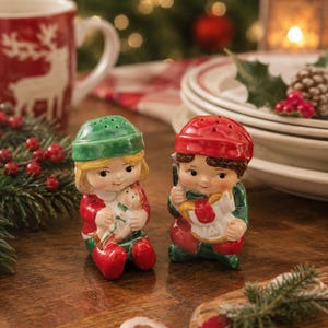 May include: A pair of ceramic salt and pepper shakers shaped like children in holiday attire. One figure wears a green hat and holds a doll, while the other wears a red hat and holds a toy horse. Both have painted facial features and colorful clothing.