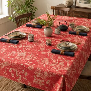 May include: A rectangular tablecloth featuring a vibrant red background with a detailed white floral and botanical pattern. The table is set with multiple ceramic bowls, small cups, dark blue cloth napkins with woven rings, and a black teapot.