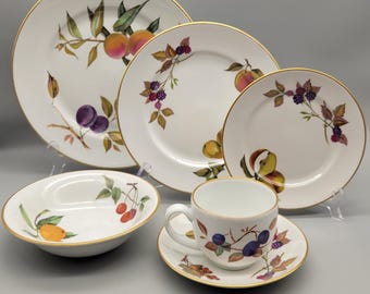 May include: A white porcelain dinnerware set featuring a fruit design with peaches, plums, and berries. The set includes plates, a bowl, a teacup, and a saucer, all with gold rims. The fruit illustrations are in shades of red, purple, and yellow.