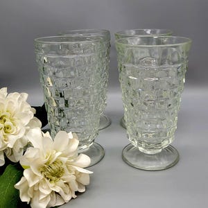 May include: Four clear, vintage-style drinking glasses with a textured, grid-like pattern. The glasses have a pedestal base and a tall, slightly flared shape. White flowers and green leaves are in the foreground, set against a gray background.