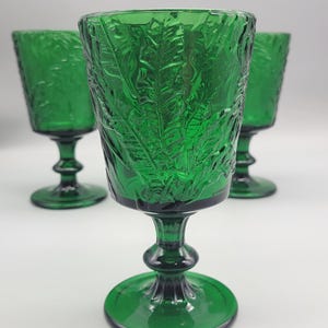 May include: Three green stemmed glasses with a leaf pattern. The glasses have a round base, a detailed stem, and a textured bowl. The glasses are made of a transparent green glass. The glasses are suitable for serving beverages.
