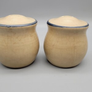 May include: Two ceramic salt and pepper shakers. The shakers are beige with a blue rim around the lid. The shakers have a bulbous shape and a small opening on top for dispensing salt and pepper. The shakers are on a light gray background.