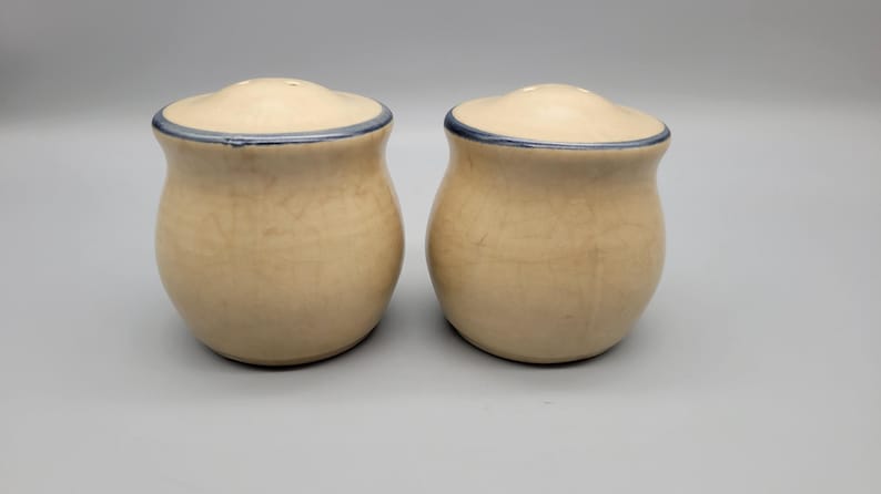 May include: Two ceramic salt and pepper shakers. The shakers are beige with a blue rim around the lid. The shakers have a bulbous shape and a small opening on top for dispensing salt and pepper. The shakers are on a light gray background.