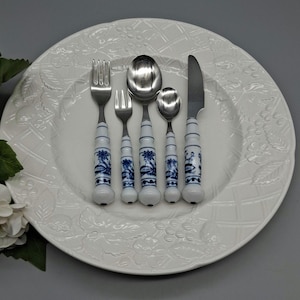 May include: A set of five pieces of silverware, including a fork, knife, and three spoons, arranged on a white plate. The silverware features white handles with blue floral designs. The plate has a raised floral pattern.