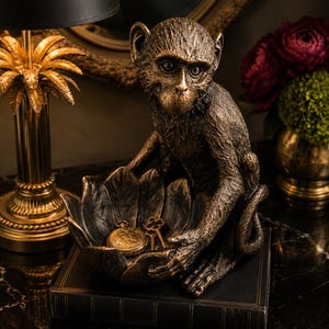 May include: A bronze-colored monkey figurine holding a bowl shaped like a large leaf. Inside the leaf bowl are a gold-toned pocket watch and two small keys. The monkey has textured fur and is sitting on top of a dark, rectangular book.