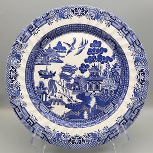 May include: A round ceramic plate featuring a blue and white willow pattern. The design depicts a landscape with a pagoda, a bridge, a boat, and two birds in flight. The rim is decorated with a geometric border and floral motifs in blue.