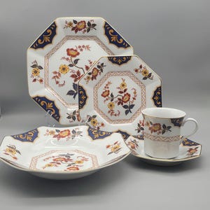 May include: A white porcelain dinnerware set with a floral design. The set includes plates, a bowl, a cup, and a saucer. The plates and bowl have a unique octagonal shape with a gold trim and navy blue accents. The floral pattern features red and yellow flowers.