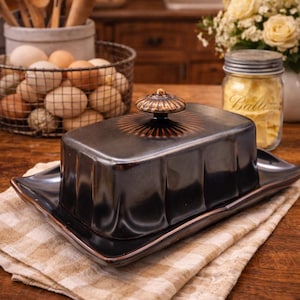 May include: A black ceramic butter dish with a lid and a decorative handle. The dish sits on a matching tray, placed on a tan and white checkered cloth. The kitchen setting includes eggs, wooden spoons, and flowers.
