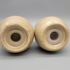 May include: Two beige ceramic salt and pepper shakers, viewed from the bottom. Each shaker has a round base with a central hole and a plastic insert. The shakers are egg-shaped and have a glossy finish.
