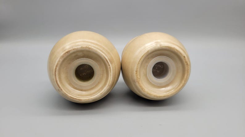 May include: Two beige ceramic salt and pepper shakers, viewed from the bottom. Each shaker has a round base with a central hole and a plastic insert. The shakers are egg-shaped and have a glossy finish.