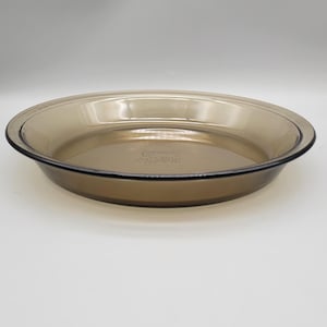 May include: A clear, round, amber-tinted Pyrex pie dish. The dish has a slightly raised rim and a smooth, transparent surface. The Pyrex logo is visible on the bottom of the dish. Ideal for baking pies and quiches.