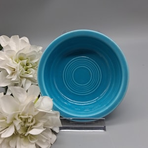 May include: A turquoise ceramic bowl with concentric circles in the center. The bowl is set on a clear acrylic stand, with white flowers in the background. The bowl has a glossy finish.