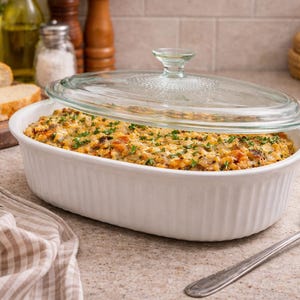 May include: An oval white ceramic baking dish with vertical ribbed sides, filled with a golden-brown baked casserole topped with green herbs. The dish is covered by a clear, circular glass lid with a textured knob handle and a decorative rim.