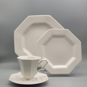 May include: A white octagonal dish set, including a teacup, saucer, and two plates. The plates and saucer have a decorative border. The teacup has a unique handle design. The set is displayed on a light gray background.