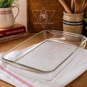 May include: A rectangular clear glass baking dish sits on a white and red striped towel. The dish is empty and has a simple, classic design. The background includes cookbooks, a ceramic pitcher, and wooden utensils.