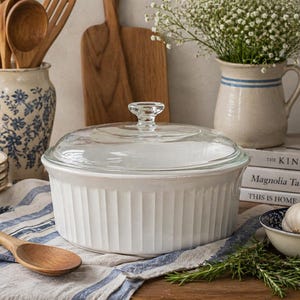 May include: A white ceramic casserole dish with a glass lid. The dish has vertical ribbed detailing and a clear glass knob. A wooden spoon and other kitchenware are in the background.