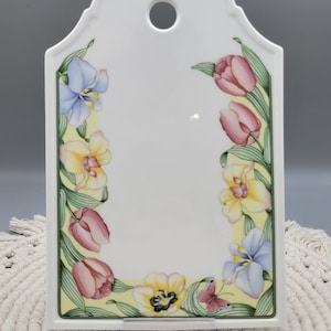 May include: White ceramic wall plaque with a floral design. The plaque features a border of pink tulips, yellow orchids, and blue irises. A small butterfly is also included in the design. The plaque has a hole at the top for hanging.