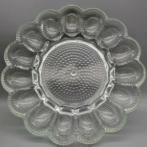May include: Clear glass egg serving platter with a scalloped edge and 12 egg-shaped indentations. The center of the platter has a textured, circular design. A decorative piece for serving eggs.