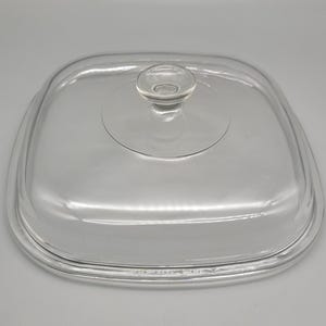 May include: A clear, square glass lid with a rounded knob handle. The lid is slightly curved and has a raised edge. The glass is transparent, allowing a clear view of the contents below. A kitchenware item.