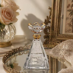 May include: A clear glass perfume bottle with a pyramid shape and a gold-colored floral design on the top. The bottle has etched floral patterns and sits on a gold-rimmed mirrored tray, with a bouquet of roses in the background.