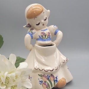 May include: A vintage ceramic figurine of a girl in a Dutch outfit. The figurine is cream-colored with painted details, including a brown hairstyle, blue accents, and floral patterns. The figurine is approximately 6 inches tall.