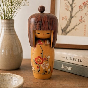 May include: A wooden Kokeshi doll with a brown, fringed hairstyle and a small, round head. The doll's body is yellow with a floral design in red and white. The doll is a traditional Japanese craft.