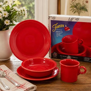 May include: A scarlet red Fiesta dinnerware set including a dinner plate, a bowl, and a mug, displayed with its original cardboard packaging box. The box features the Fiesta logo, product details, and images of the included red ceramic dishware pieces.