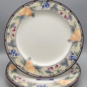 May include: A stack of three white ceramic dinner plates. Each plate features a decorative border with a fruit and vine design, including peaches, grapes, and leaves. The plates have a brown rim and a cream-colored background.