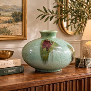 May include: A wide, low-profile ceramic vase with a textured, light green glaze. The surface features a prominent, dark reddish-brown abstract splash pattern on the front, with a smooth, rounded body and a short, narrow neck opening.