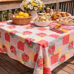 May include: A picnic table set for a meal, featuring a floral patterned tablecloth with red roses, yellow squares, and blue accents. A basket of lemons, a vase of daisies, and plates of food are arranged on the table.