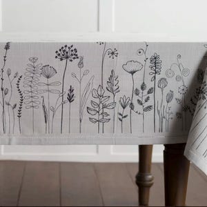 May include: A light gray tablecloth with a black floral design. The design features various stylized flowers and plants along the border. The tablecloth is draped over a dark wooden table.
